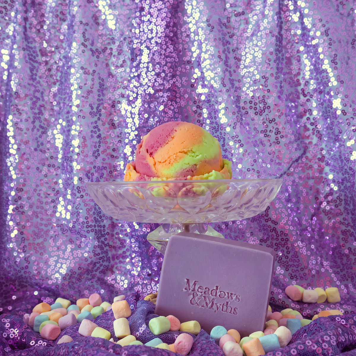 Meadows & Myths 'Glitch' purple rectangle soap with 'Meadows & Myths' embossed on it. Sitting on purple sequins fabric with the same fabric draped in the background. The raised dish has a scoop of rainbow sherbet. Colorful marshmallows are scattered on the ground.  