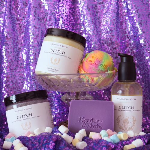 A collection of Meadows & Myths Glitch body care featuring a jar of body butter, jar of sugar scrub, purple rectangle soap, and bottle of body oil. Sitting on purple sequins fabric with the same fabric draped in the background. The body butter sits in a glass raised dish with a scoop of rainbow sherbet. Colorful marshmallows are scattered on the ground.  