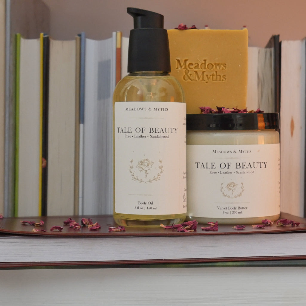Meadows & Myths Tale of Beauty body care set featuring a jar of body butter, bottle of oil, yellow rectangle soap. Styled on a stack of books with dried rose petals scattered around it and a row of books in the background.