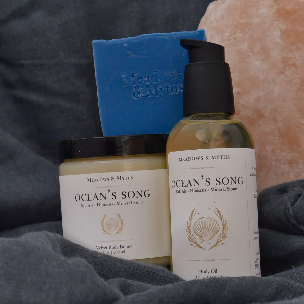 Meadows & Myths 'Ocean's Song' scented body care set featuring a blue rectangular soap bar, a bottle of body oil, and a jar of body butter, styled with a pink salt stone and dark gray-blue fabric.