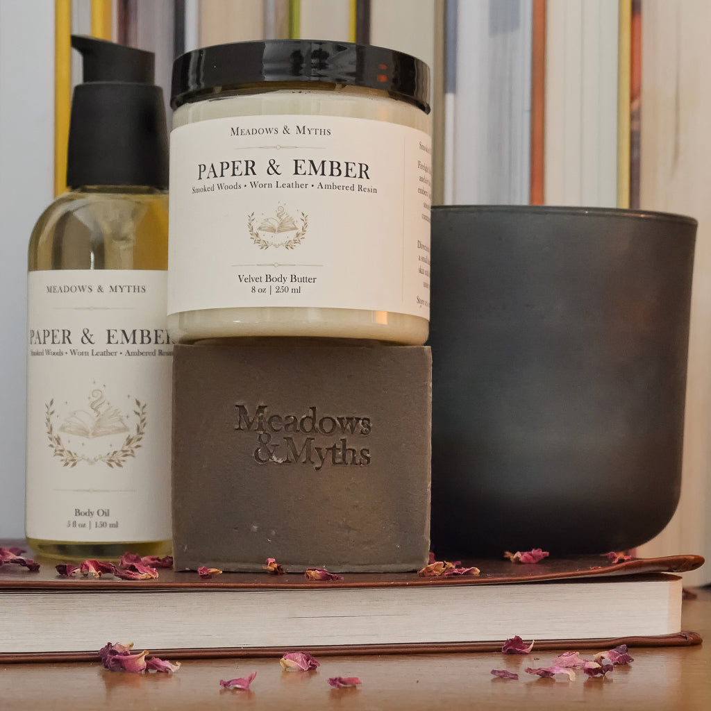 Meadows & Myths Paper & Ember body care set featuring a jar of body butter, bottle of oil, brown rectangle soap. Styled on a wooden surface with dried rose petals scattered around it. They sit on top of a closed leather bound book, with a dark green candle and a row of books in the background.