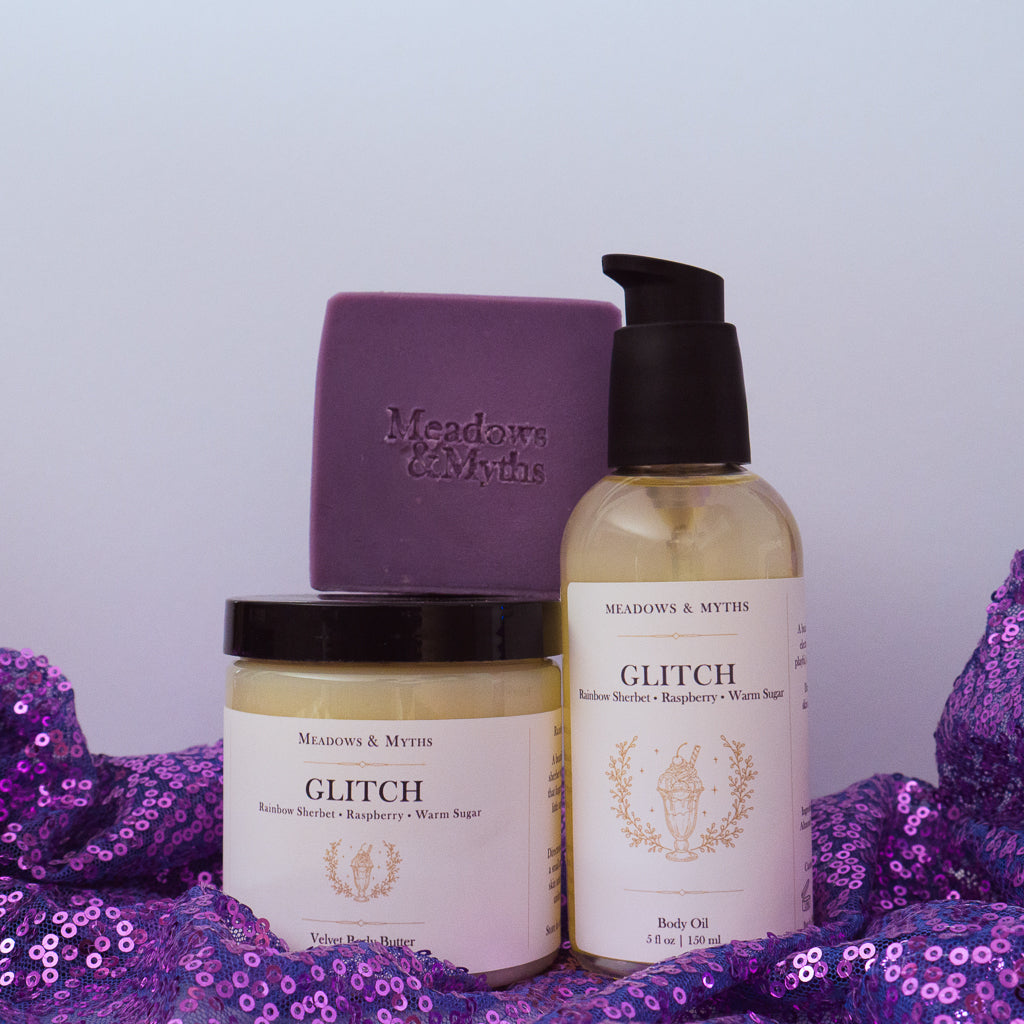 Meadows & Myths 'Glitch' scented body care set featuring a purple rectangular soap bar with "Meadows & Myths" embossed on it, a bottle of body oil, and a jar of body butter, sitting on purple sequins fabric.