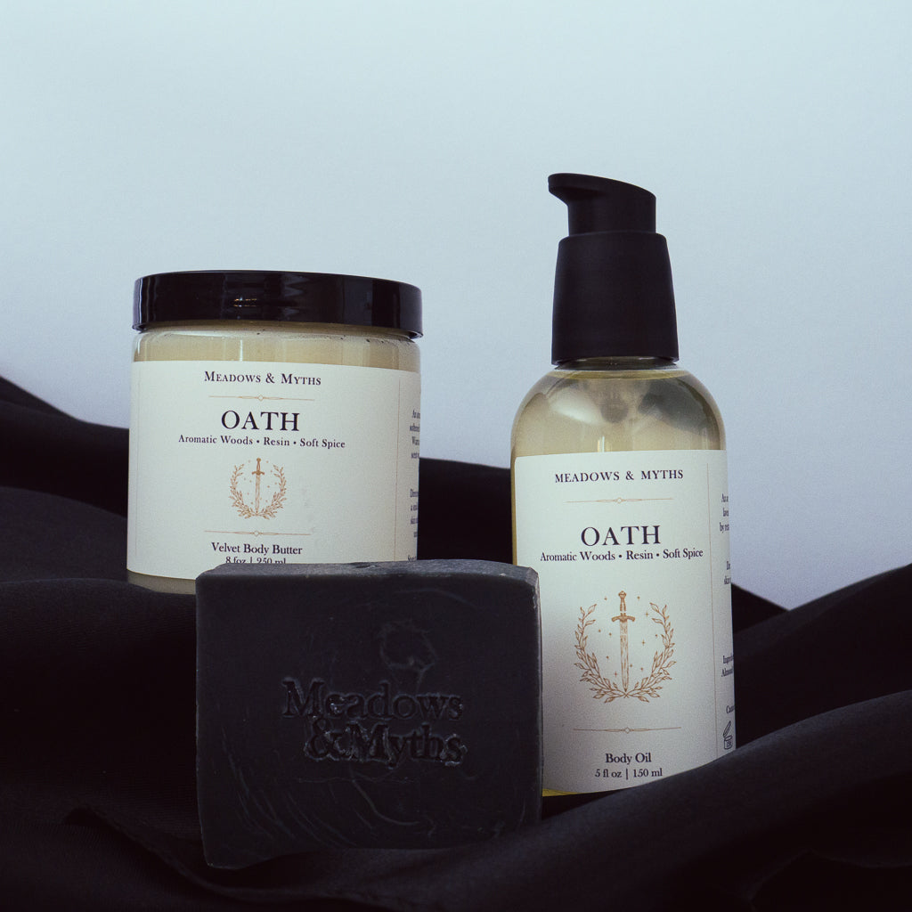 Meadows & Myths 'Oath' scented body care set featuring a black rectangular soap bar with "Meadows & Myths" embossed on it, a bottle of body oil, and a jar of body butter, sitting on black fabric.