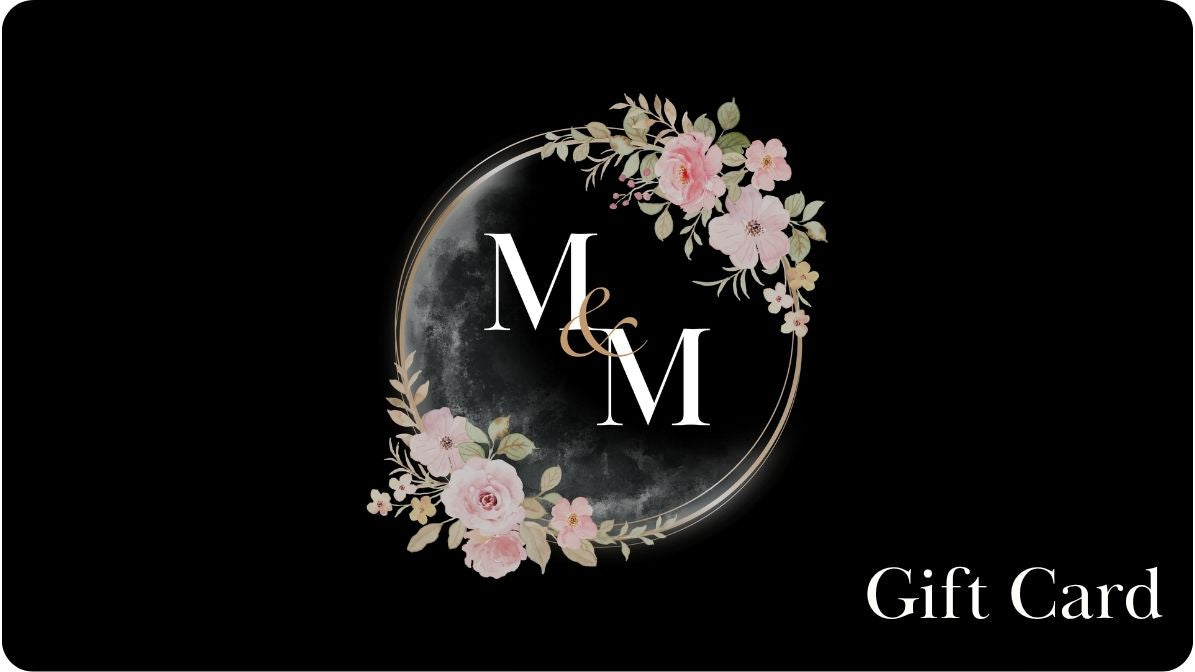 Gift card with Meadows & Myths logo. Gift card is black with white writing. Logo is a circle with multiple thin gold outlines of circles. Pink flowers with light green leaves on the lower left and upper right side. M & M , in white, is in the middle. A faded black and white crescent moon sits on the right side. 