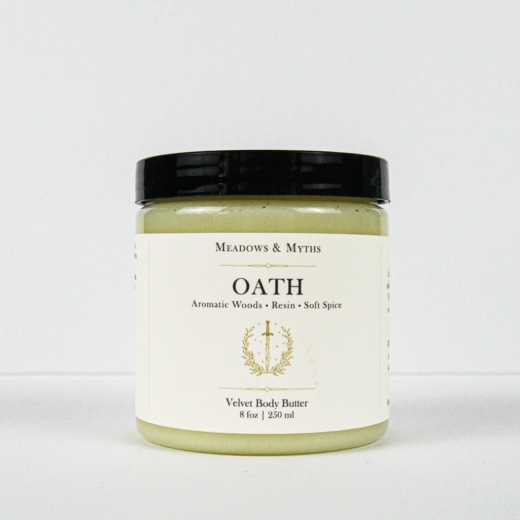 A jar of Meadows & Myth's Oath Velvet Body Butter, featuring a light greenish-yellow balm visible through the clear jar, a cream-colored label with gold text, and a black lid, set against a plain white background.