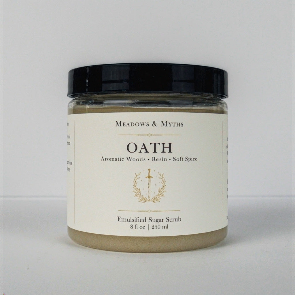 A jar of Meadows & Myth's 'Oath' emulsified sugar scrub, featuring a cream-colored label with gold detailing and a black lid, set against a plain light gray background.