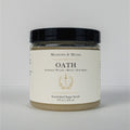 A jar of Meadows & Myth's 'Oath' emulsified sugar scrub, featuring a cream-colored label with gold detailing and a black lid, set against a plain light gray background.