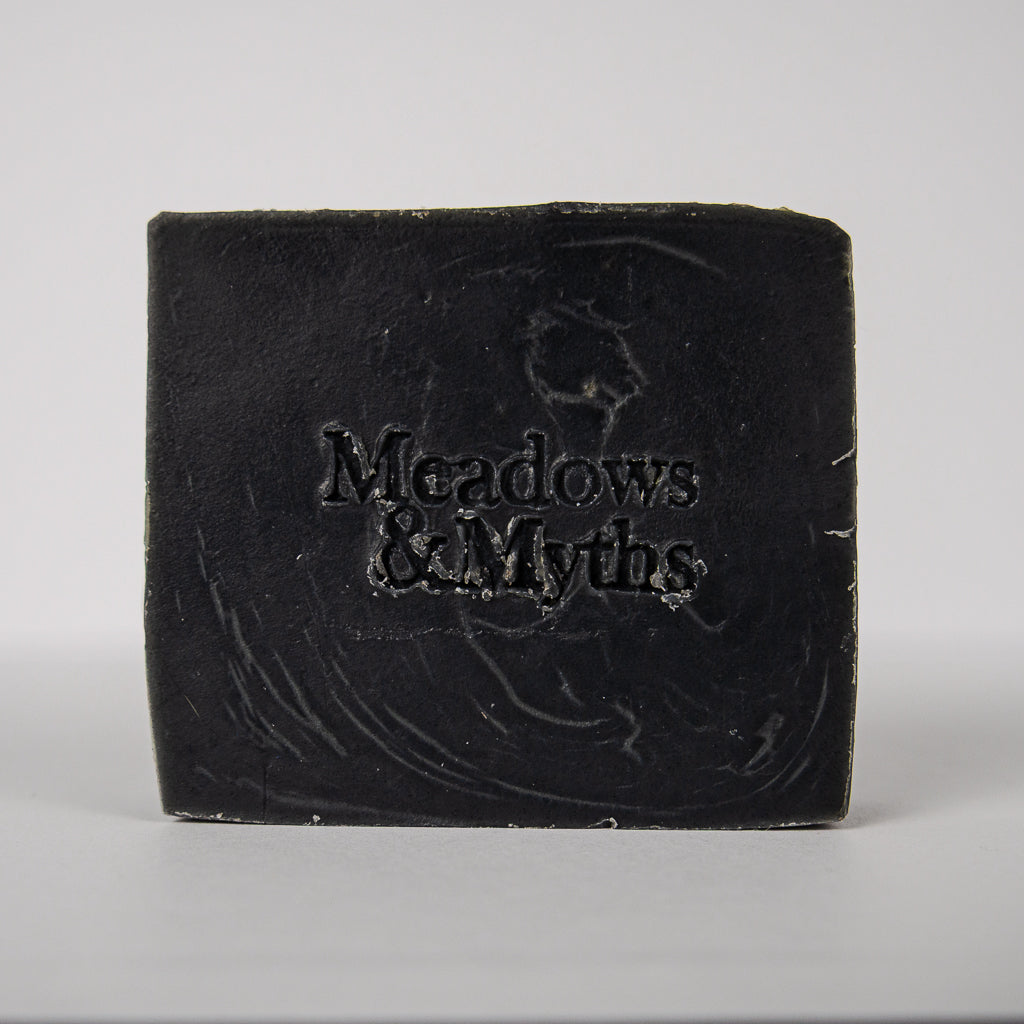 A rectangular bar of dark soap with the brand name 'Meadows & Myths' embossed on the front, set against a plain light gray background.