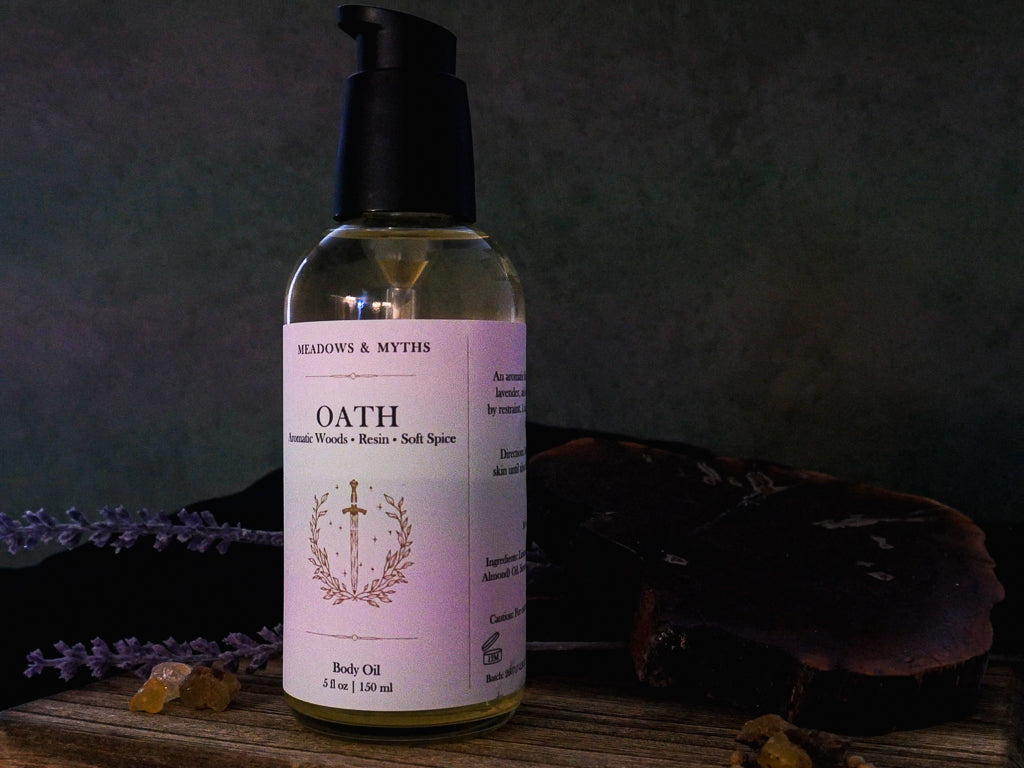 A bottle of Meadows & Myths 'Oath' body oil with a black pump and a cream label, on a rustic wooden surface with dried lavender, resin, and a dark background.