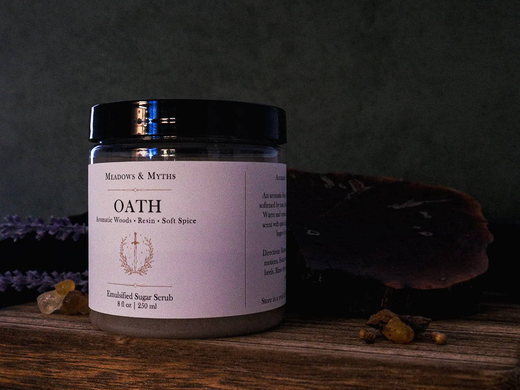 A jar of 'Oath' emulsified sugar scrub by 'Meadows & Myths' on a rustic wooden surface with dried lavender, resin, and a dark background.