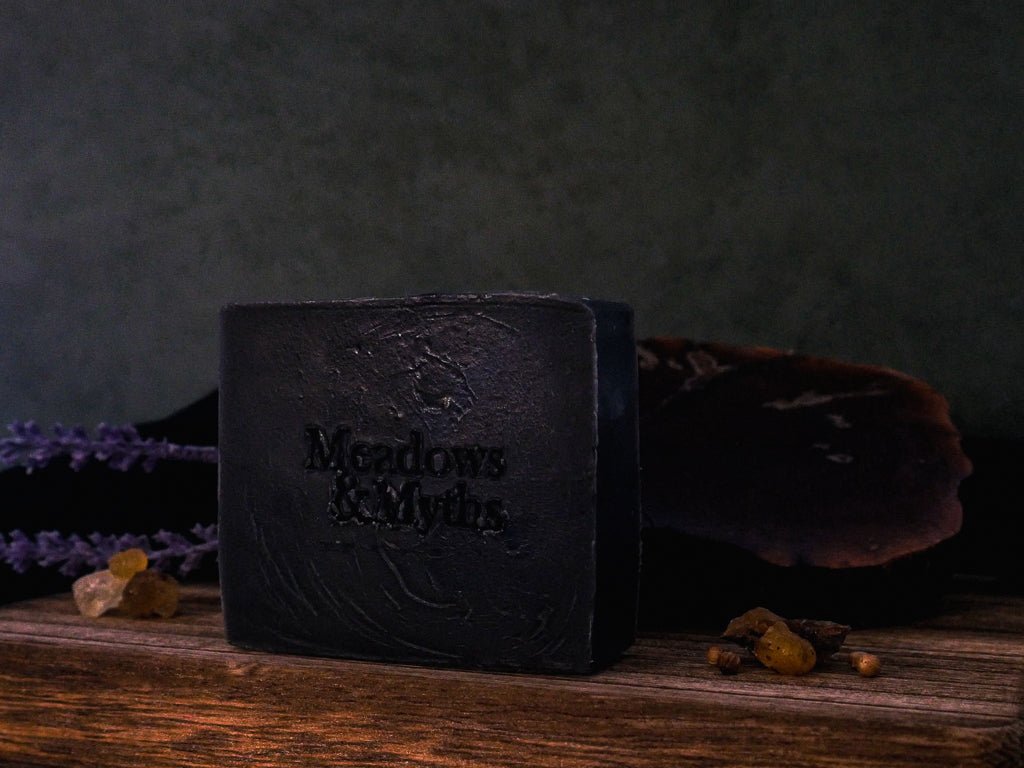 A dark charcoal-colored rectangular bar of soap with the brand name 'Meadows & Myths' embossed on the front, on a rustic wooden surface with dried lavender, resin, and a dark background.