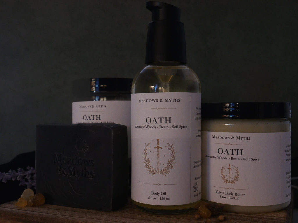 A collection of Meadows & Myths natural products including a body oil bottle, body butter jar, soap bar, and body sugar scrub jar, on a rustic wooden surface with dried lavender, resin, and a dark background.