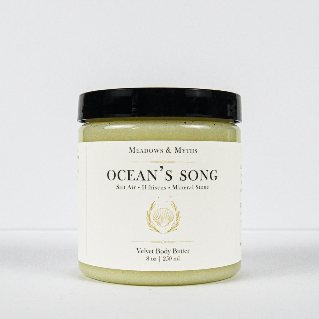 A jar of Meadows & Myth's 'Ocean's Song' velvet body butter, featuring a white label with gold detailing and a black lid, set against a plain white background.