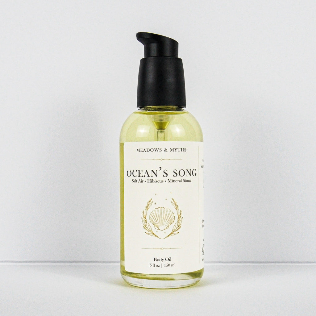 A bottle of Meadows & Myth's 'Ocean's Song' body oil, featuring a clear glass bottle filled with pale yellow oil, a black pump dispenser, and a cream-colored label with gold detailing of a shell, set against a plain white background.