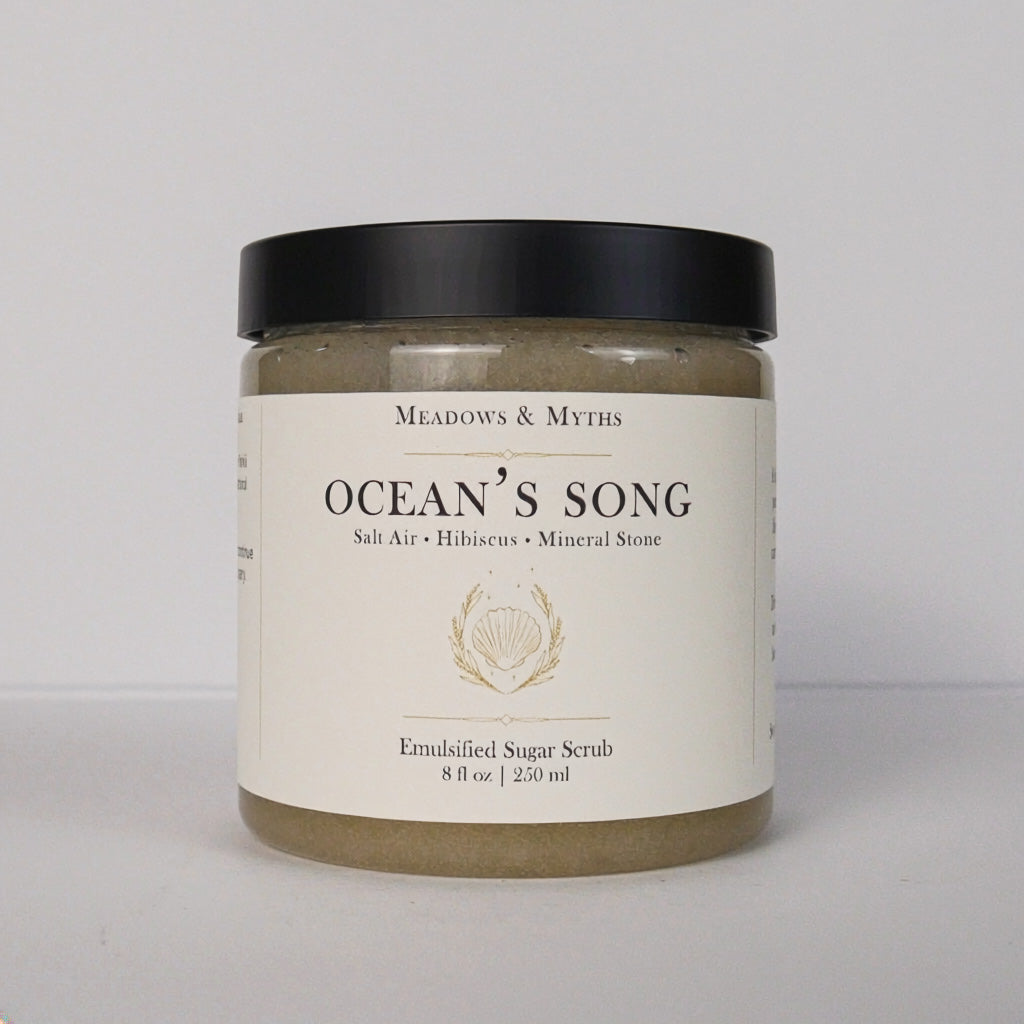 A jar of Meadows & Myth's 'Ocean's Song' emulsified sugar scrub, featuring a black lid and a cream-colored label with gold detailing, set against a plain light gray background.