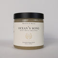 A jar of Meadows & Myth's 'Ocean's Song' emulsified sugar scrub, featuring a black lid and a cream-colored label with gold detailing, set against a plain light gray background.