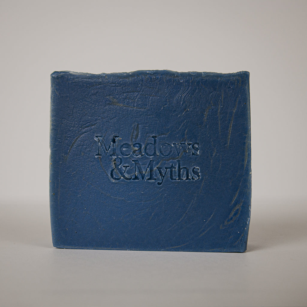 A rectangular blue bar of soap standing upright on a light gray background, embossed with the brand name 'Meadows & Myths'.
