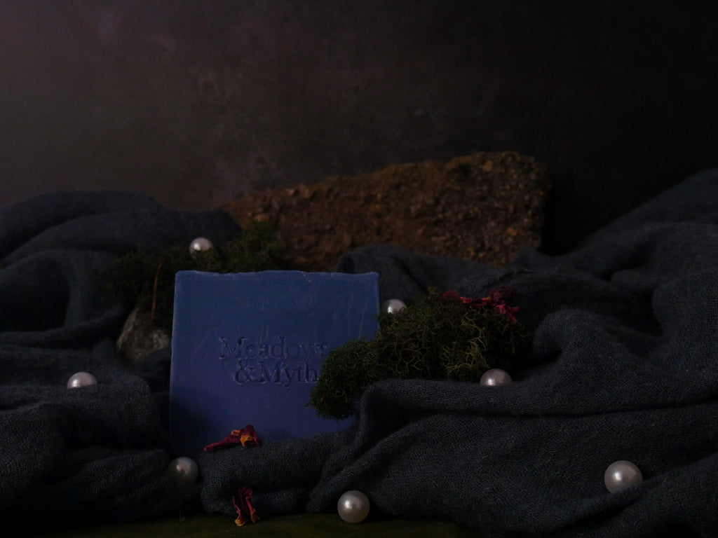 A rectangular blue soap bar with embossed text reading 'Meadow & Myths' sits on a dark charcoal draped fabric, surrounded by faux moss, white faux pearls, and small dried red flowers against a dark textured background.