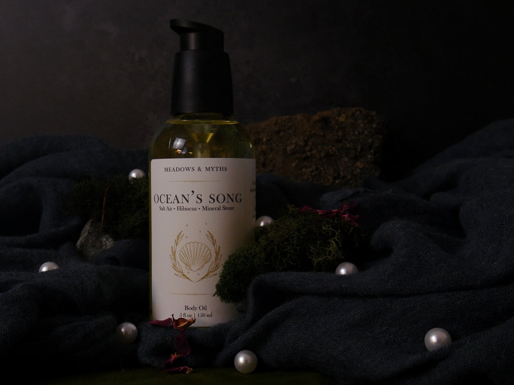A bottle of 'Ocean's Song' body oil by Meadows & Myths, featuring a black pump and a cream-colored label, styled on a dark draped fabric with moss, pearls, and dried flowers.