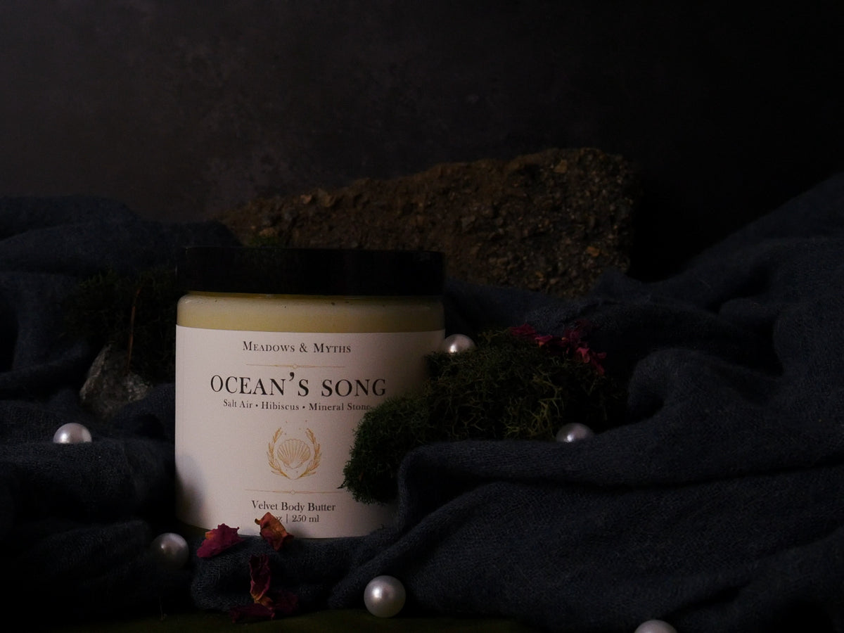 A jar of Velvet Body Butter labeled 'Ocean's Song' by Meadows & Myths, styled on dark draped fabric with moss, pearls, and dried flowers against a dark background.