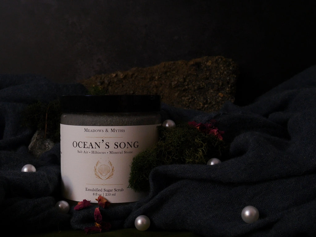 A jar of Emulsified Sugar Scrub labeled 'Ocean's Song' by Meadows & Myths, styled on dark draped fabric with moss, pearls, and dried flowers against a dark background.