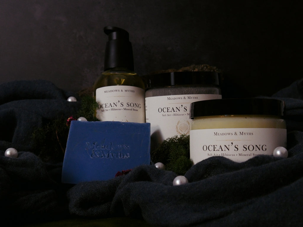 A collection of Meadows & Myths bath and body products, including a blue bar of soap, a pump bottle of Body Oil, two jars: Body Butter and Sugar Scrub, arranged on a dark textured fabric with moss and pearl accents.