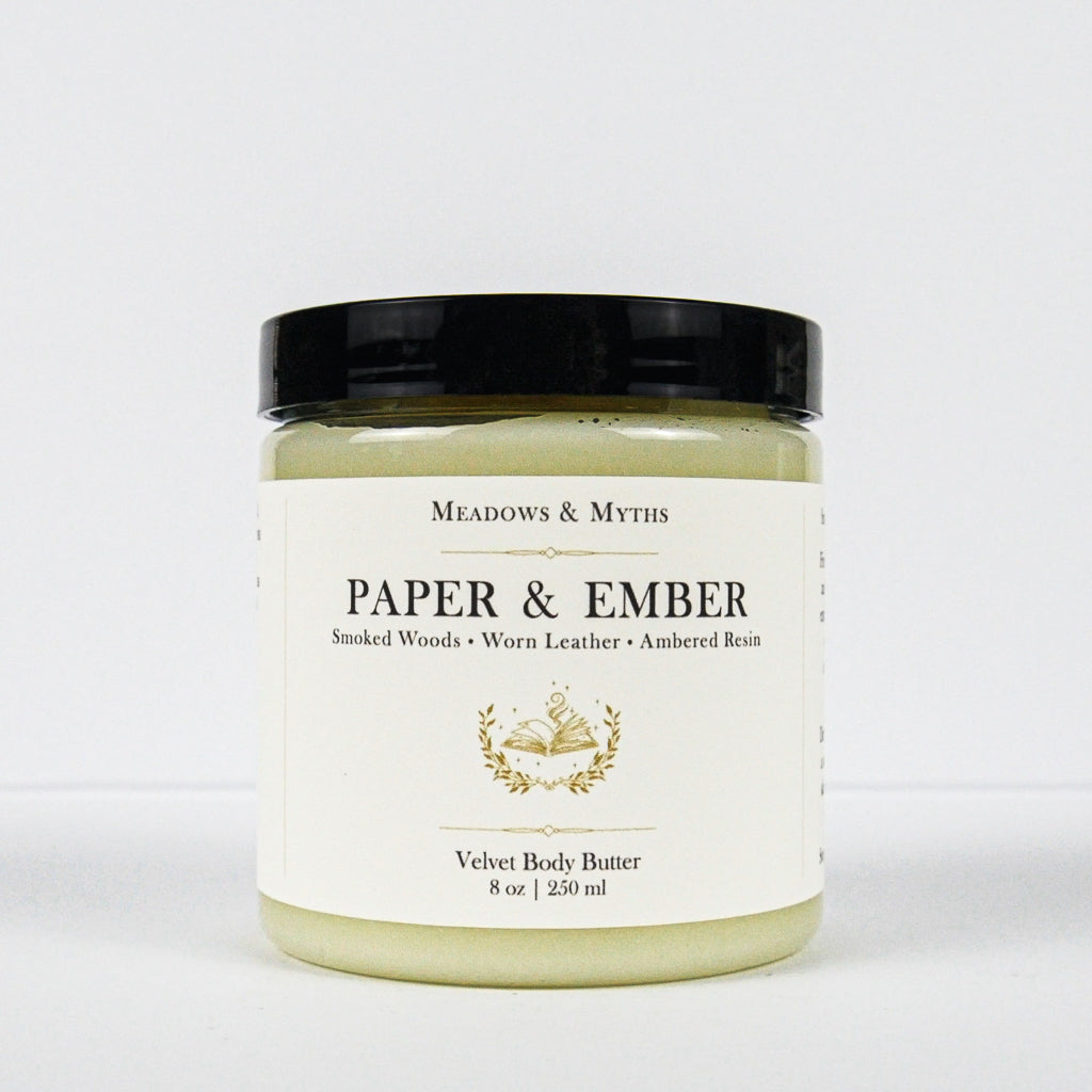 A jar of Meadows & Myth's 'Paper & Ember' velvet body butter, featuring a cream-colored label with gold detailing and a black lid, set against a plain white background.