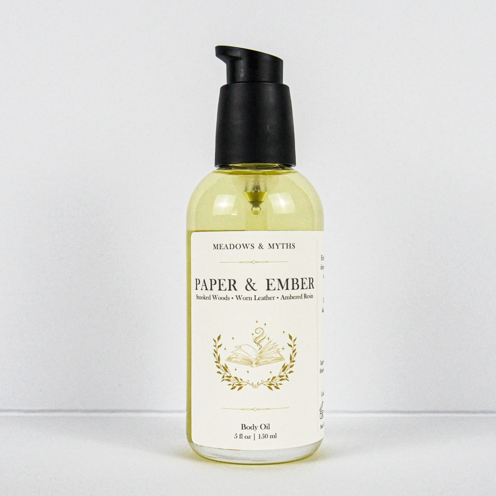A bottle of Meadows & Myth's Paper & Ember body oil, featuring a clear bottle filled with pale yellow oil, a cream-colored label with gold detailing, and a black pump dispenser, set against a plain white background.