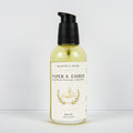 A bottle of Meadows & Myth's Paper & Ember body oil, featuring a clear bottle filled with pale yellow oil, a cream-colored label with gold detailing, and a black pump dispenser, set against a plain white background.
