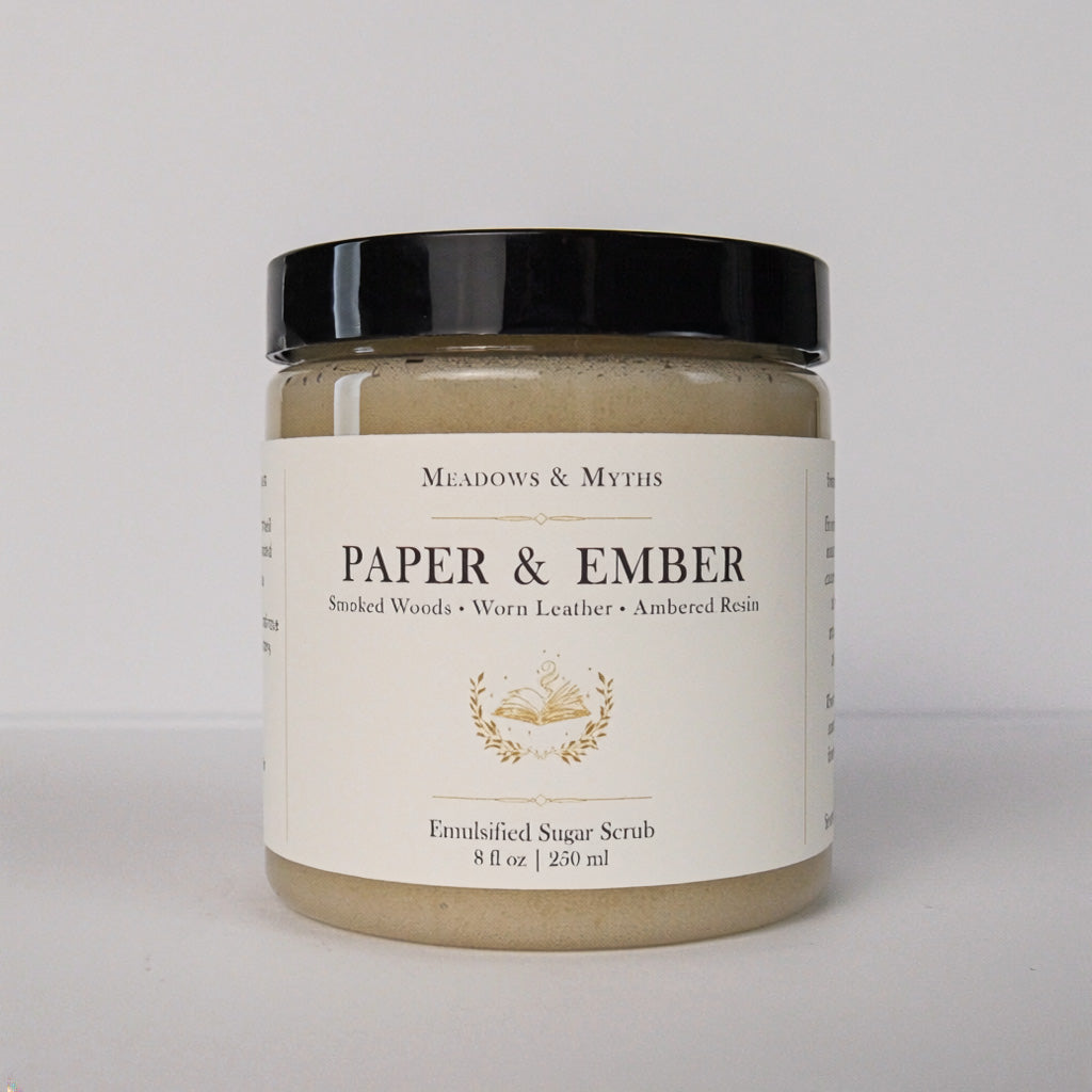 A jar of Meadows & Myth's Paper & Ember Emulsified Sugar Scrub, featuring a black lid and a cream-colored label with gold detailing, set against a plain light gray background.