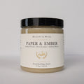 A jar of Meadows & Myth's Paper & Ember Emulsified Sugar Scrub, featuring a black lid and a cream-colored label with gold detailing, set against a plain light gray background.