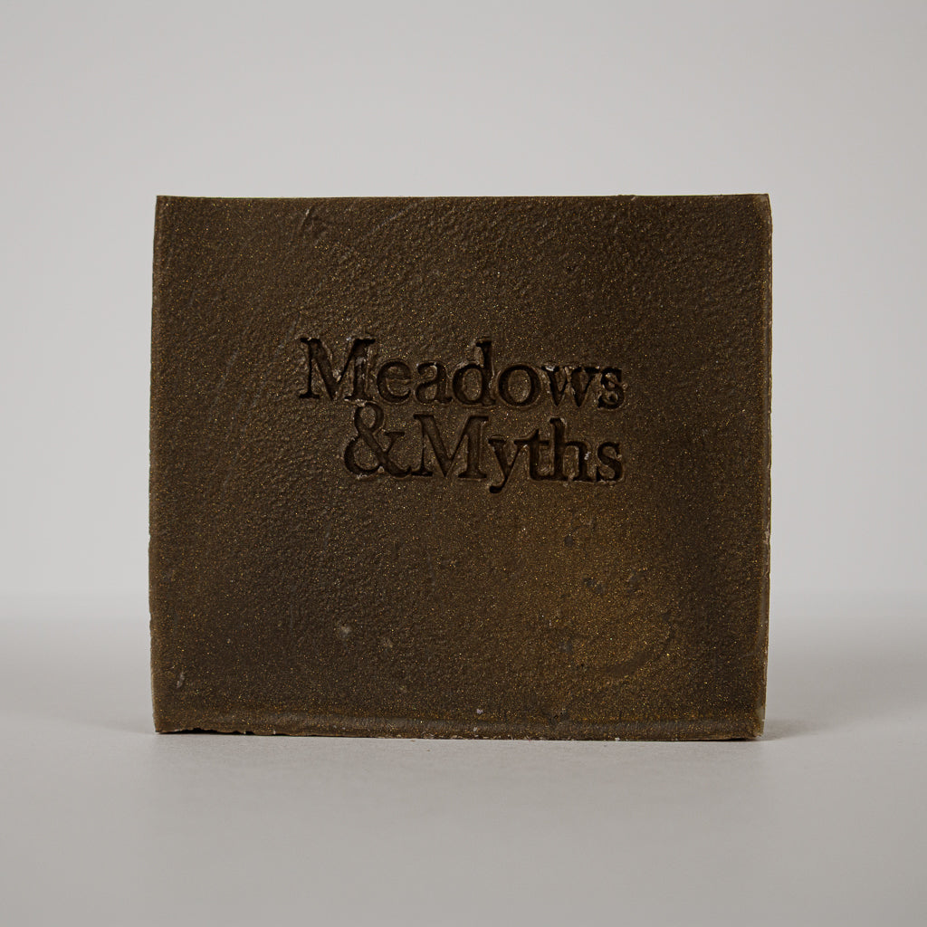 A square dark brown textured block or box with the embossed text 'Meadows & Myths' on the front, photographed against a plain light gray background.