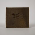 A square dark brown textured block or box with the embossed text 'Meadows & Myths' on the front, photographed against a plain light gray background.