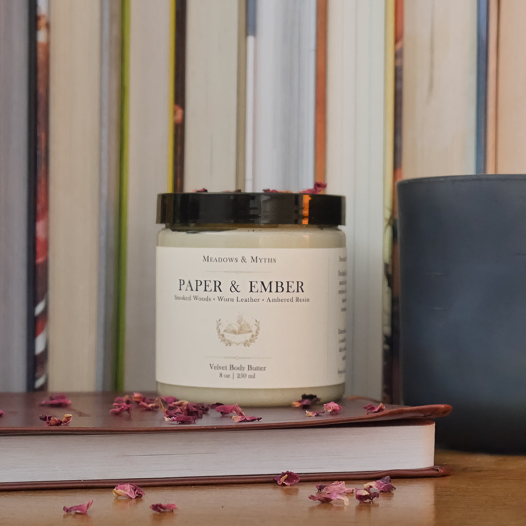 A jar of body butter labeled 'Paper & Ember' by Meadows & Myths sits on a wooden surface with dried rose petals scattered around it. The jar is styled on top of a closed leather bound book, with a dark green candle and a row of books in the background.