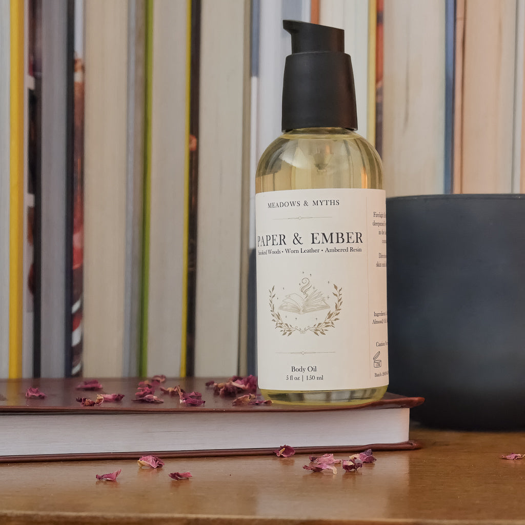 A bottle of body oil labeled 'Paper & Ember' by Meadows & Myths sits on a wooden surface with dried rose petals scattered around it. The bottle is styled on top of a closed leather bound book, with a dark green candle and a row of books in the background.