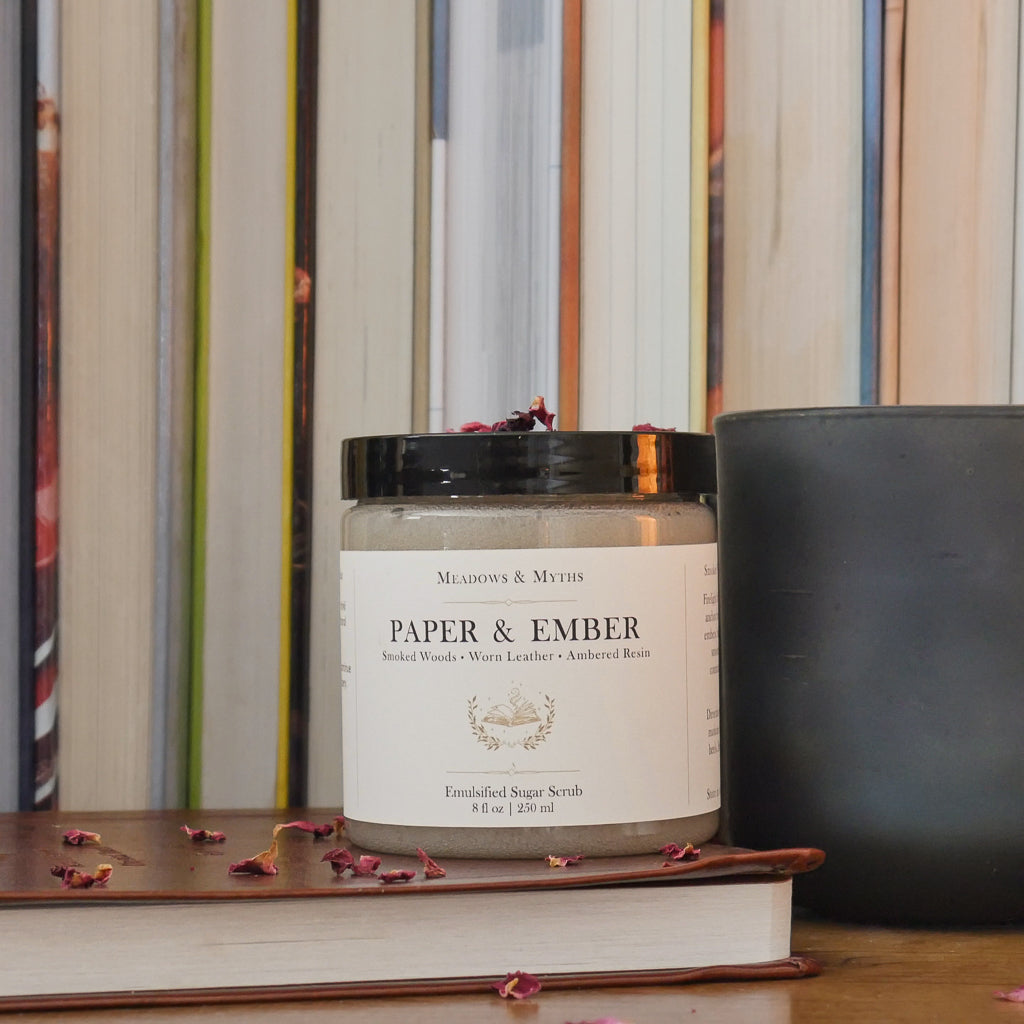 A jar of sugar scrub labeled 'Paper & Ember' by Meadows & Myths sits on a wooden surface with dried rose petals scattered around it. The jar is styled on top of a closed leather bound book, with a dark green candle and a row of books in the background.
