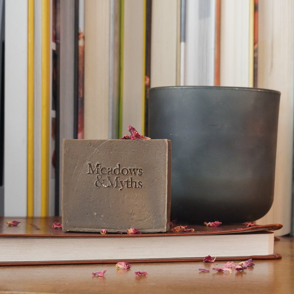 A brown rectangle soap with "Meadows & Myth" embossed on it.  labeled 'Paper & Ember' sits on a wooden surface with dried rose petals scattered around it. The soap is styled on top of a closed leather bound book, with a dark green candle and a row of books in the background.