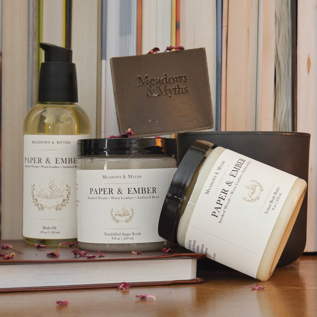 A collection of Meadows & Myths Paper & Ember body care featuring a jar of body butter, jar of sugar scrub, brown rectangle soap, and bottle of body oil. Styled on a wooden surface with dried rose petals scattered around it. They sit on top of a closed leather bound book, with a dark green candle and a row of books in the background.