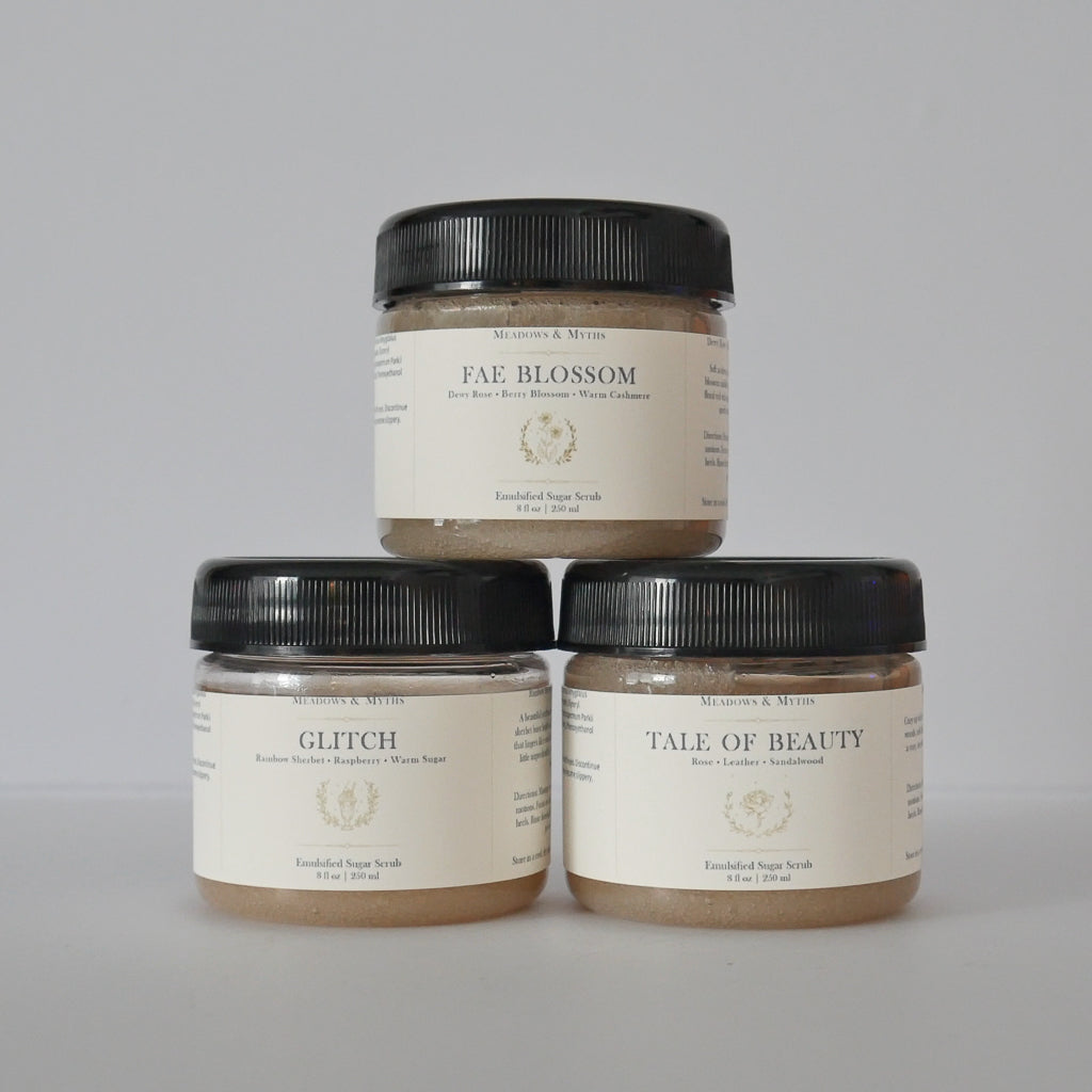 Three jars of Meadows & Myths sugar scrub products stacked in a pyramid on a light gray background. The top jar is 'Fae Blossom' and the bottom two are 'Glitch' and 'Tale of Beauty'.