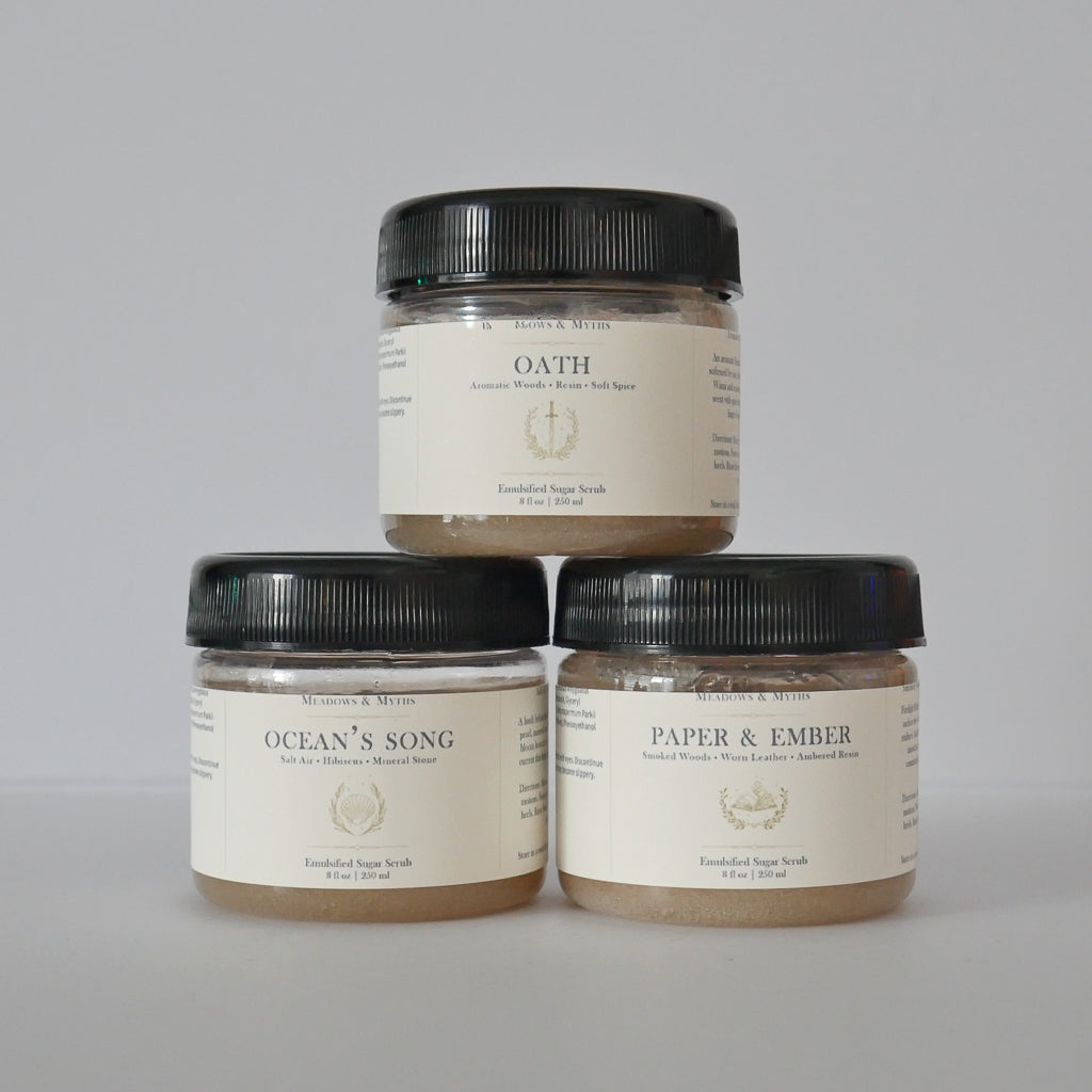 Three jars of Meadows & Myths sugar scrub products stacked in a pyramid on a light gray background. The top jar is 'Oath' and the bottom two are 'Ocean's Song' and 'Paper & Ember'.