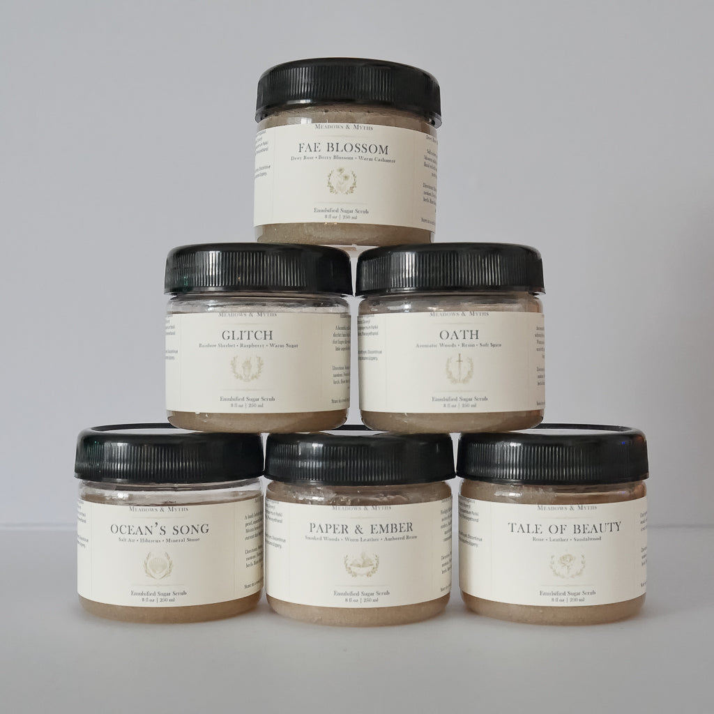 Six jars of Meadows & Myths sugar scrub arranged same jars in a pyramid stack on a light gray background. The jars have a  cream-colored label with black lids and feature labels for Tale of Beauty, Ocean's Song, Fae Blossom, Oath, Paper & Ember, and Glitch.