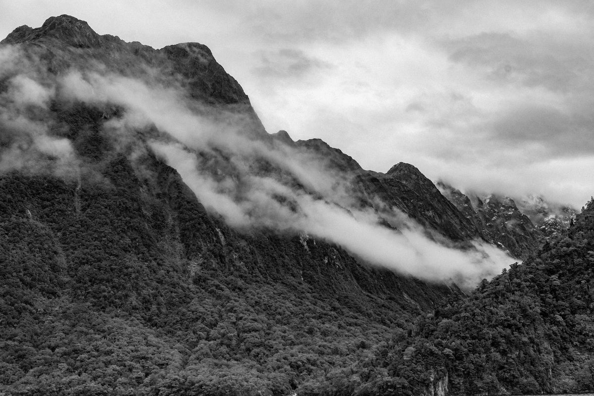 A black and white picture of tree-covered mountains with clouds rolling over the top.