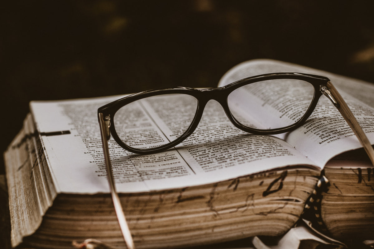 A pair of black-framed eyeglasses resting on an open, thick book with printed text.