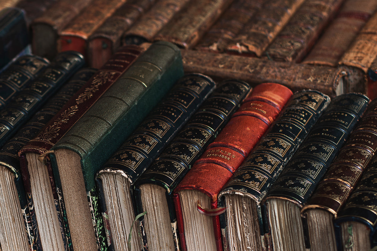 A collection of worn, antique hardcover books with ornate embossed covers, arranged closely together in rows. The books are mostly dark brown, black, and green, with one prominent red book in the center.